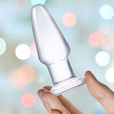 The Gläs 3.5" Glass Butt Plug, featuring a tapered design, is held between finger and thumb against a soft-focus, colorful, and festive background with bokeh lights.